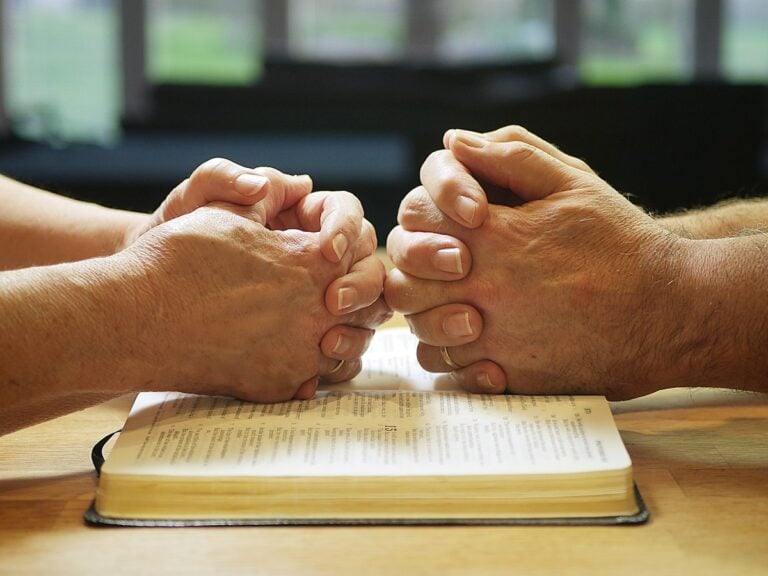 Two people praying together, hands folded over an open Bible in soft window light